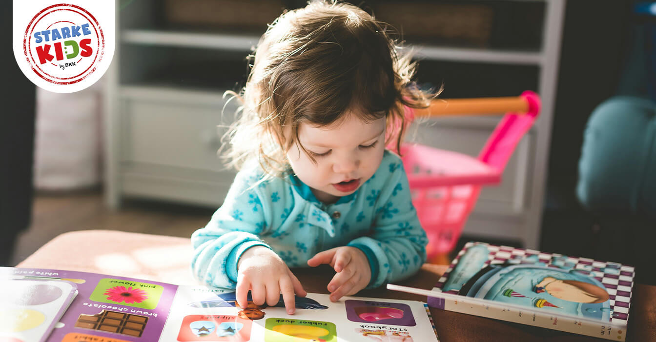 Kleinkind in blauem Schlafanzug betrachtet konzentriert ein buntes Bilderbuch im Spielzimmer mit Spielzeug im Hintergrund.