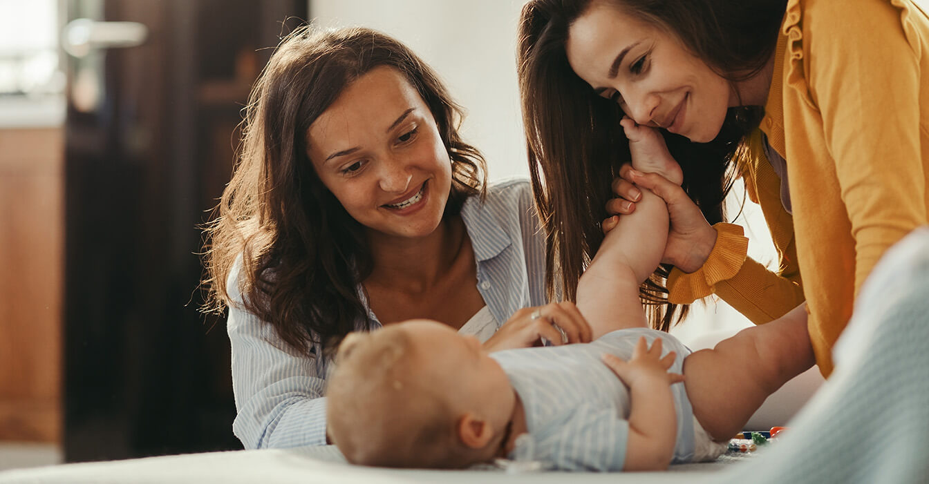 Zwei lächelnde Frauen spielen mit einem glücklichen Baby, das auf einem Bett liegt, in einem warm beleuchteten Zimmer.