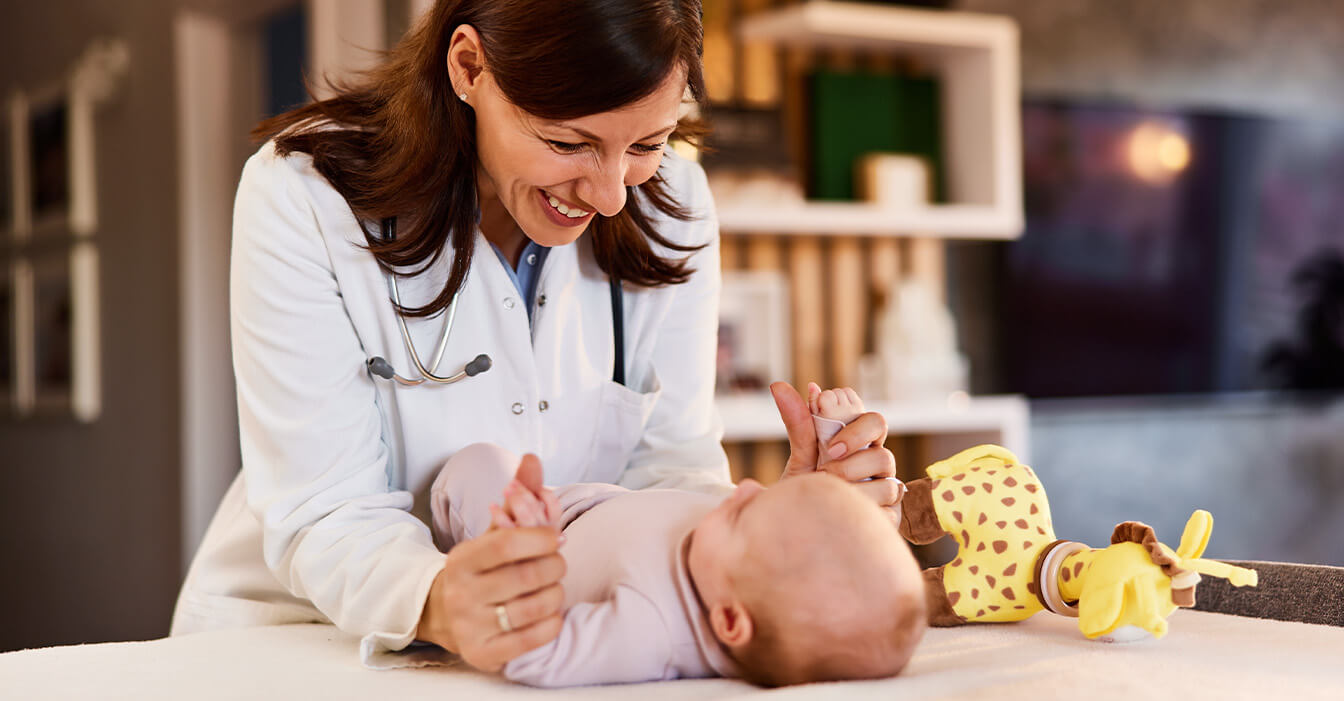 Lächelnde Ärztin in Weiß spielt mit einem glücklichen Baby auf einer Untersuchungsliege, ein Stofftier liegt neben dem Baby.