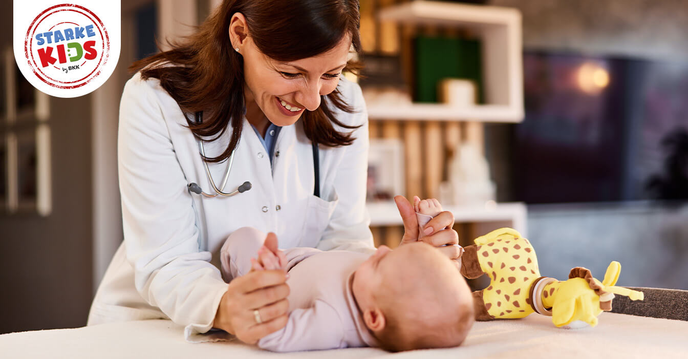 Lächelnde Ärztin in Weiß spielt mit einem glücklichen Baby auf einer Untersuchungsliege, ein Stofftier liegt neben dem Baby.