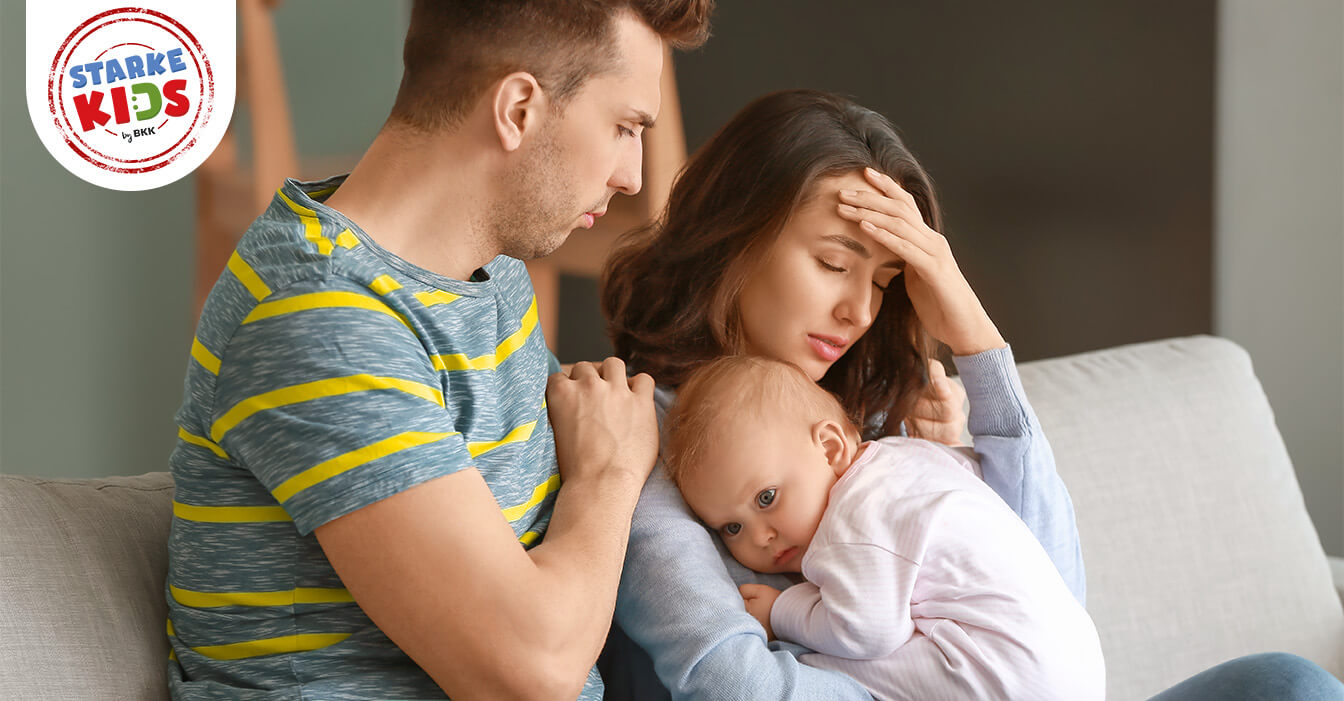 Besorgter Mann umarmt Frau, die Kopfschmerzen zu haben scheint, während sie ein kleines Kind hält, das auf ihrer Schulter ruht.