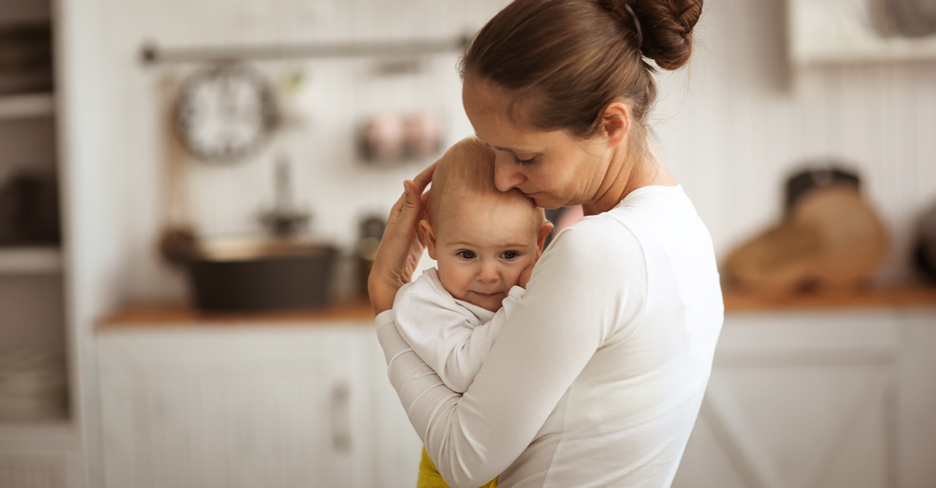 Besorgte Mutter hält ihr Baby liebevoll im Arm, in einer küchenähnlichen Umgebung.