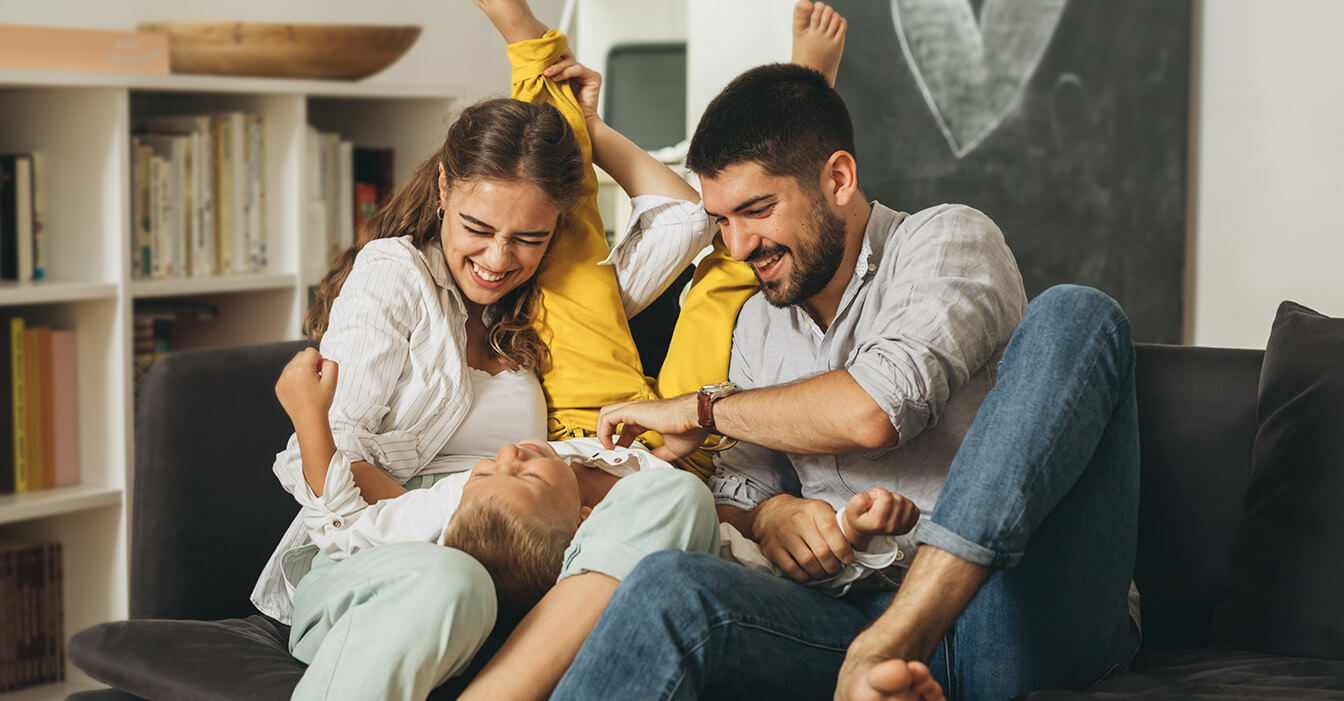Eine Familie hat Spaß zusammen auf der Couch, mit einem lachenden Kind, das auf dem Rücken liegt, umgeben von seinen fröhlich aussehenden Eltern.