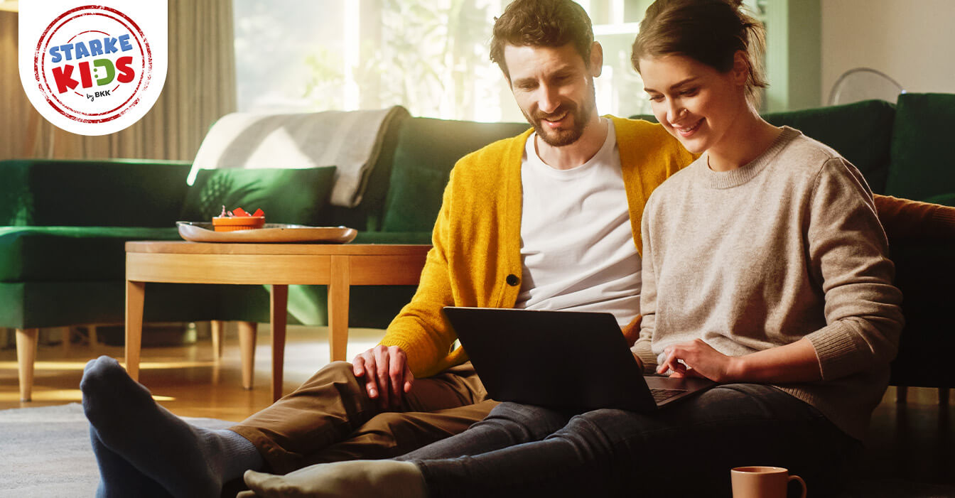 Ein Paar sitzt entspannt auf dem Boden, der Mann in einem gelben Cardigan und die Frau in einem grauen Pullover, während sie gemeinsam auf einen Laptop schauen.