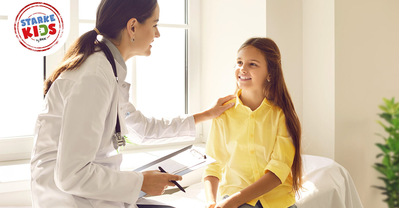 Eine freundliche Ärztin spricht mit einem lächelnden Mädchen in einem gelben Hemd während einer Konsultation in einem hellen Raum.