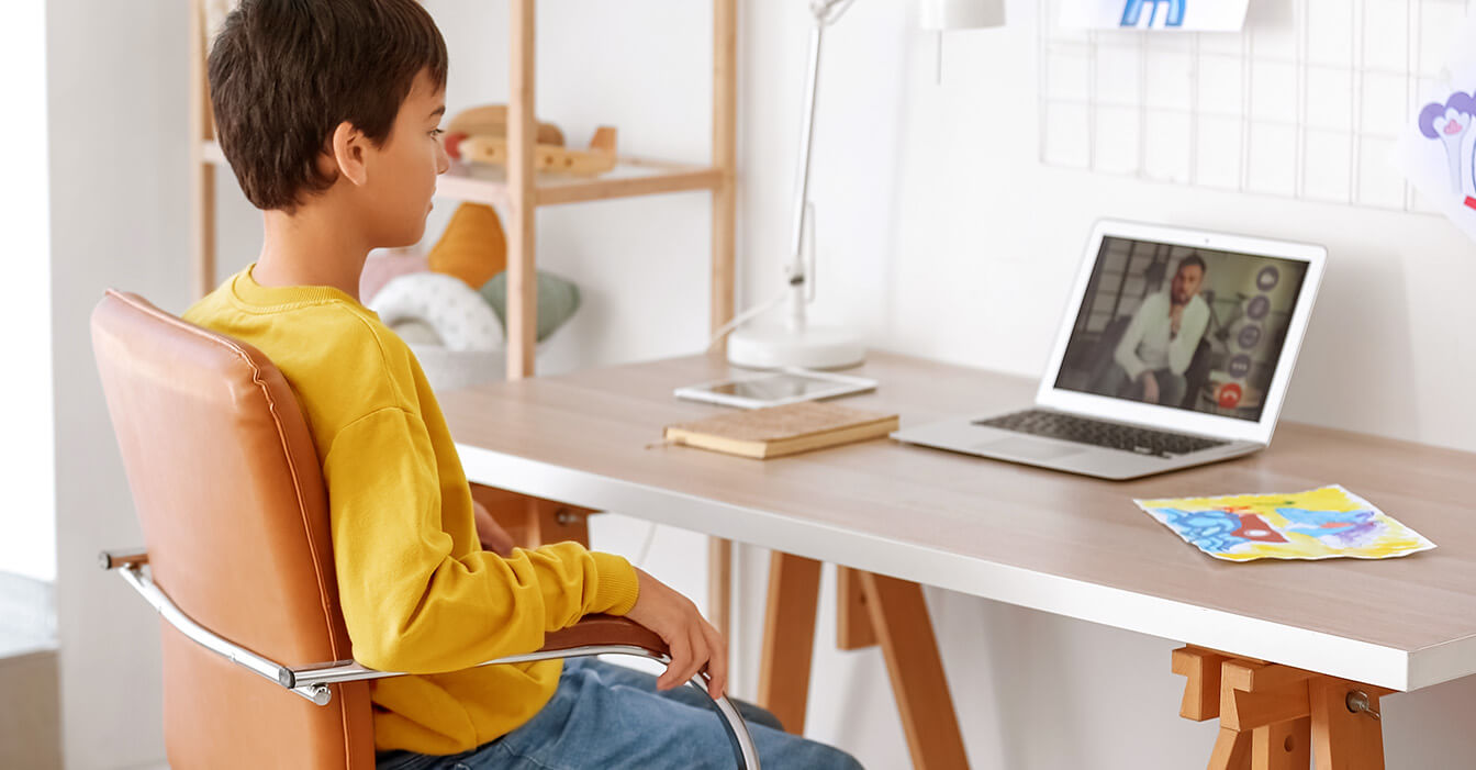 Ein Junge in einem gelben Pullover nimmt an einer Online-Sitzung mit einem Therapeuten auf seinem Laptop teil, während er an einem hellen Schreibtisch in einem kinderfreundlich eingerichteten Raum sitzt.