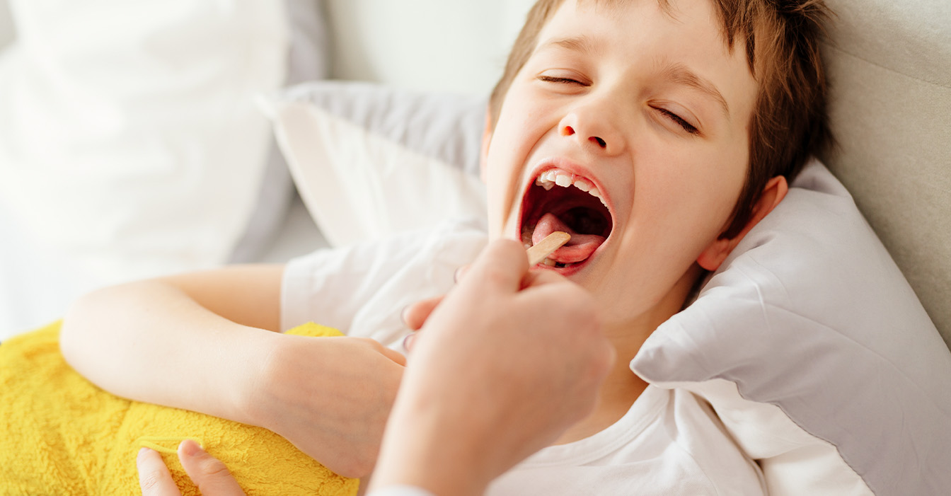Ein Junge liegt im Bett und öffnet weit den Mund für eine medizinische Untersuchung, während jemand mit einem Spatel die Zunge herunterdrückt.