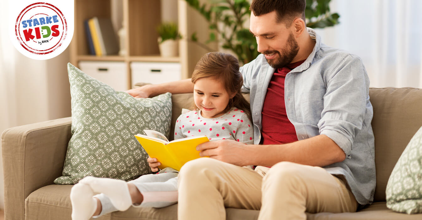 Ein lächelnder Vater liest ein Buch mit seiner Tochter, die begeistert zuhört, während sie gemütlich auf dem Sofa sitzen.