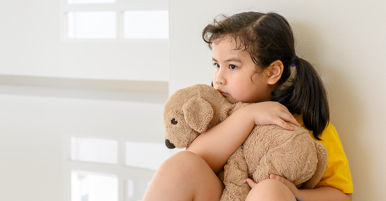 Nachdenkliches kleines Mädchen in einem gelben Shirt umarmt tröstend ihren Teddybären und sitzt auf dem Boden gegen eine Wand gelehnt.