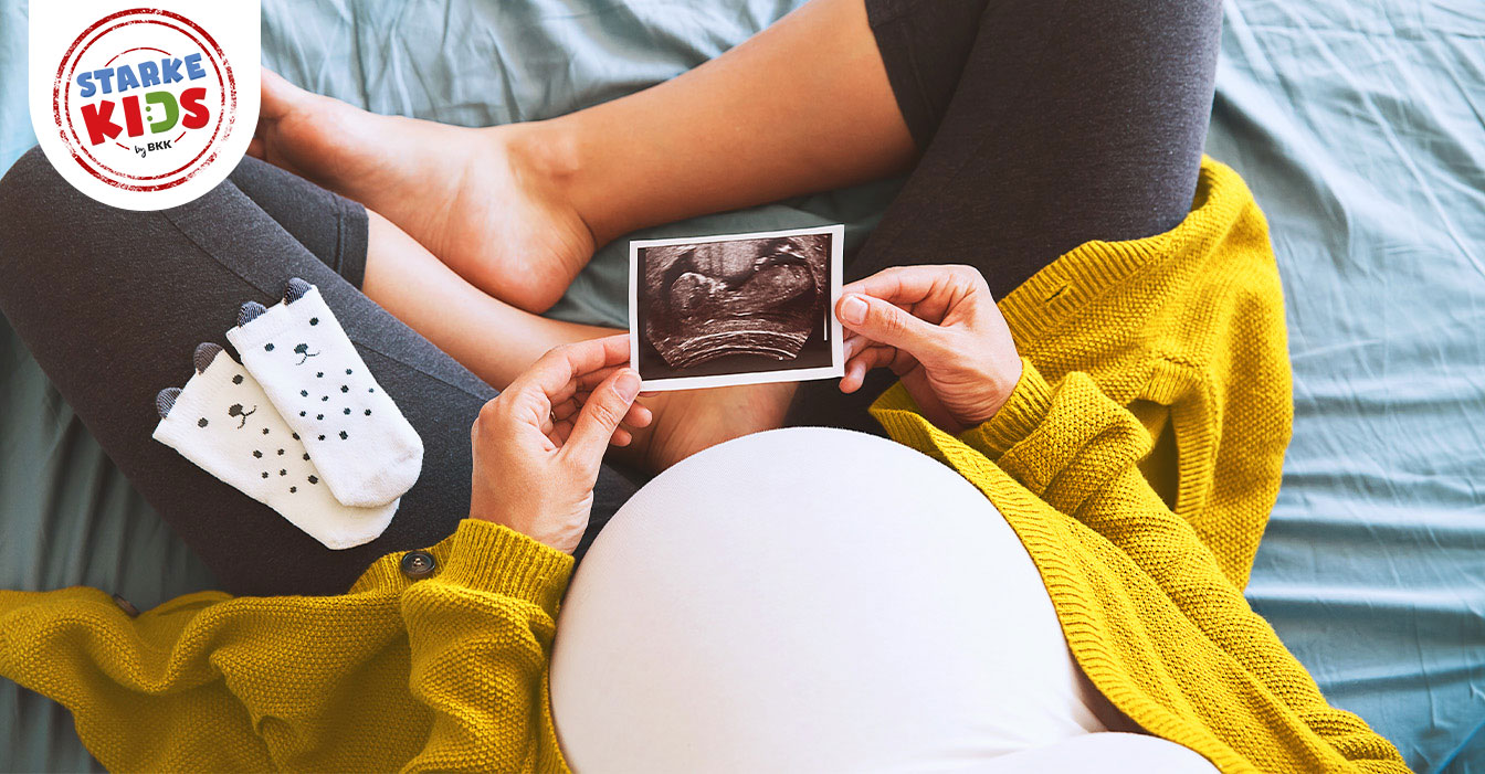 Schwangere Frau in gelber Strickjacke und grauer Leggings hält Ultraschallbild ihres Babys, während sie auf einem Bett sitzt.