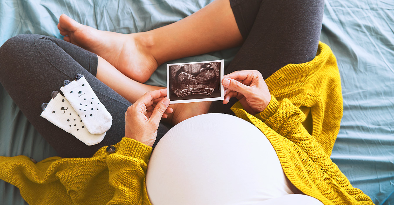 Schwangere Frau in gelber Strickjacke und grauer Leggings hält Ultraschallbild ihres Babys, während sie auf einem Bett sitzt.