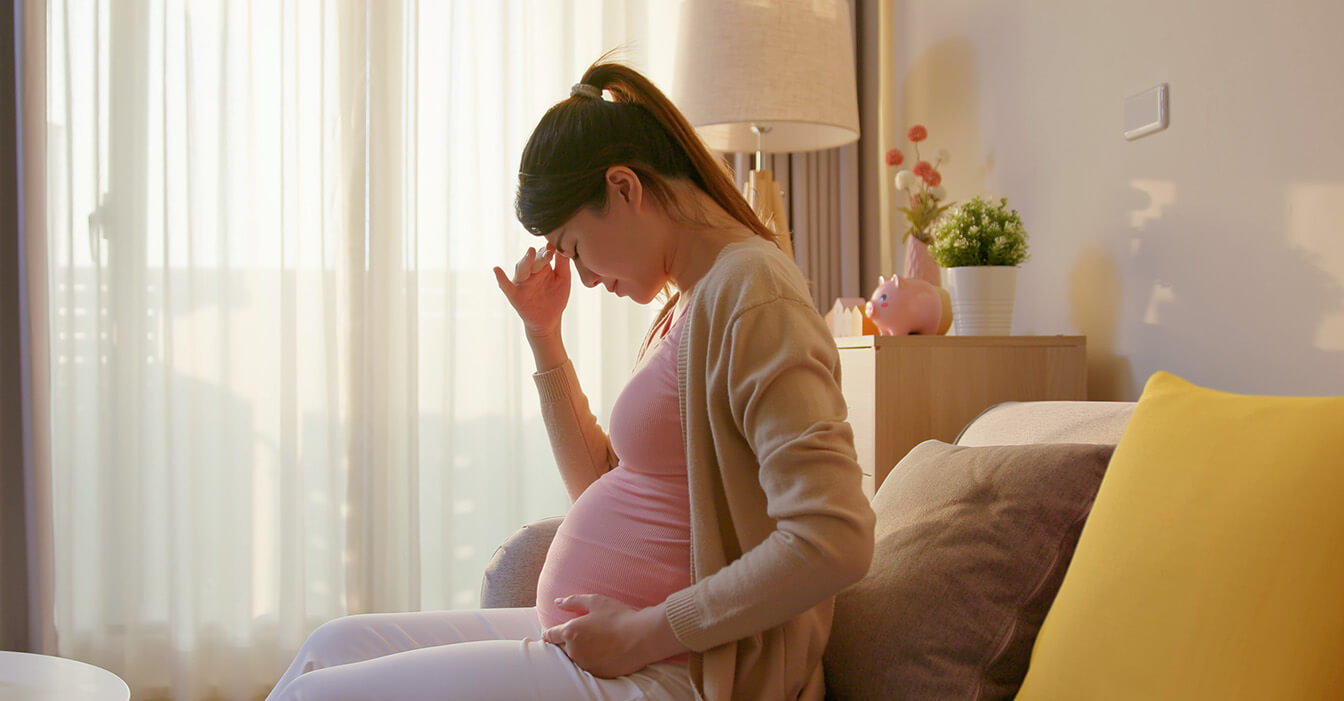 Schwangere Frau mit Kopfschmerzen sitzt auf dem Sofa, hält ihren Bauch und lehnt sich zurück, im sonnigen Zimmer mit weichen Farben.