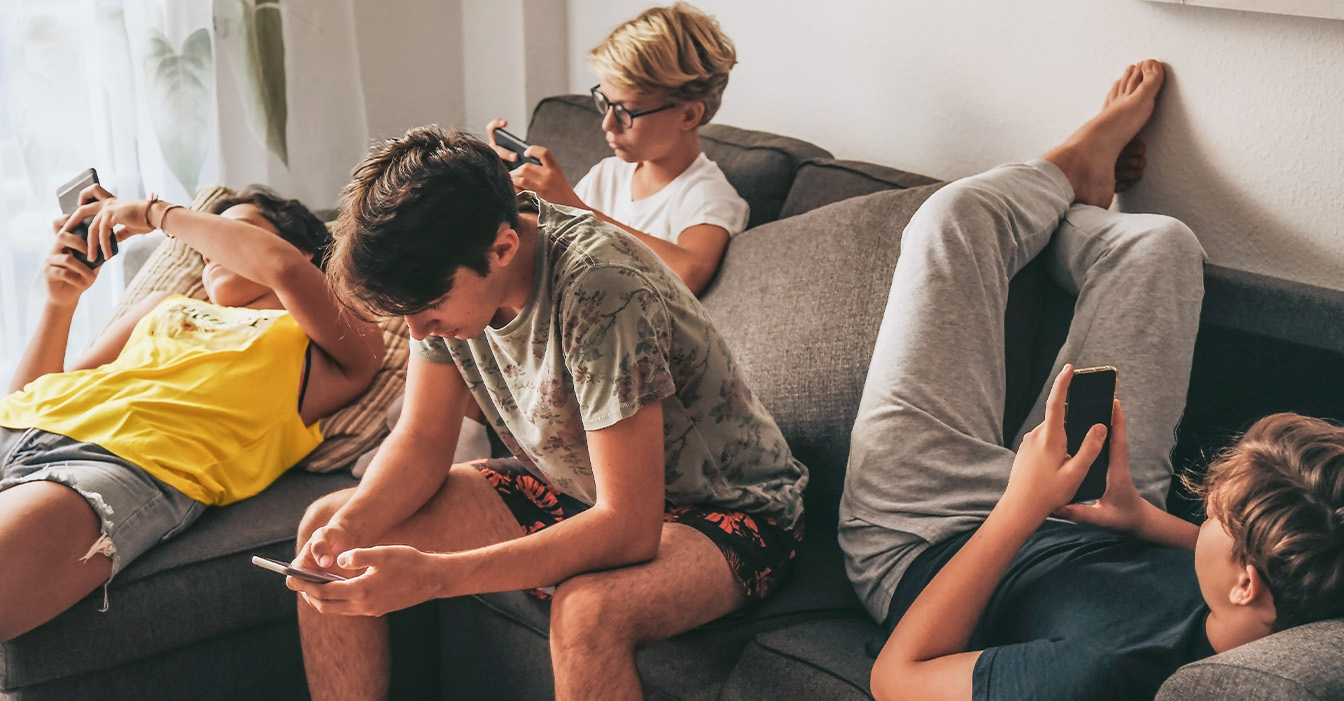 Eine Gruppe von Juggendlichen sitzt zusammen auf einem Sofa, jeder vertieft in sein eigenes Smartphone, ohne miteinander zu interagieren.