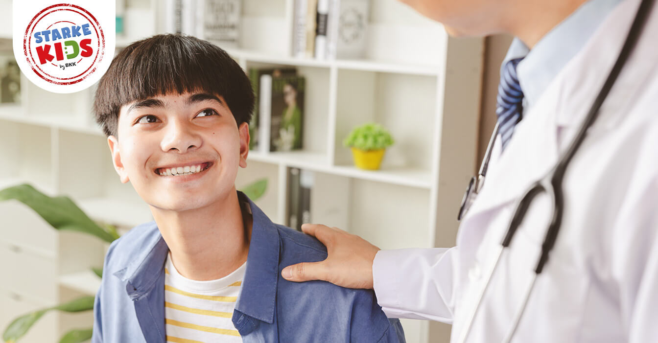 Ein lächelnder Jugendlicher schaut aufwärts zu einem Arzt, der seine Hand auf die Schulter des Jugendlichen legt.