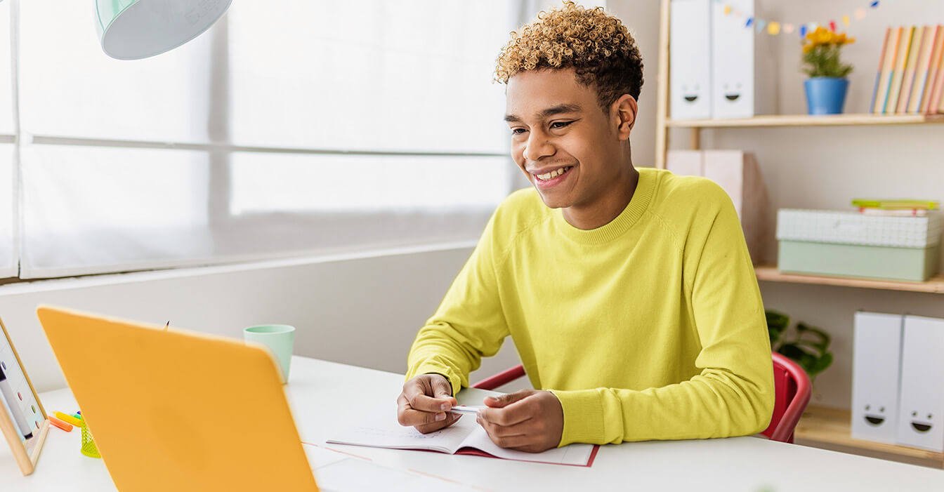 Ein fröhlicher Teenager mit lockigem Haar und einem gelben Pullover sitzt an einem Schreibtisch, lächelt und schreibt etwas, während er auf einen Laptop-Bildschirm schaut.